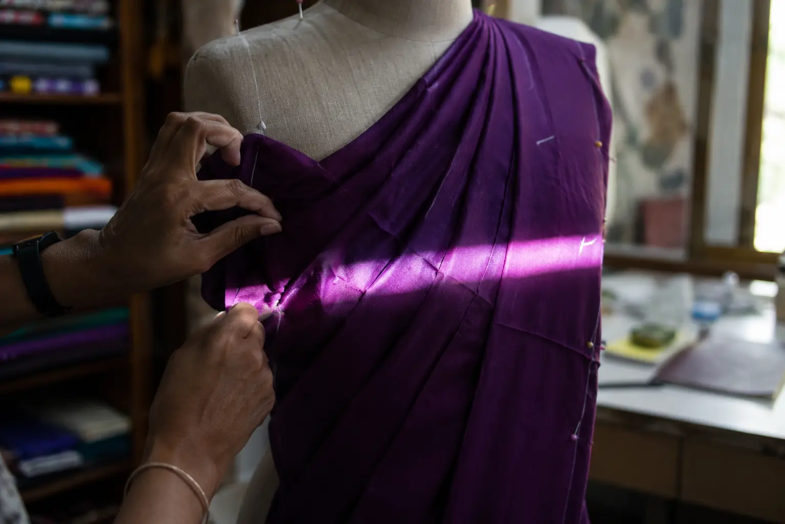 A designer's hands draping original purple fabric on a plain dress form as a narrow beam of light crosses the folds