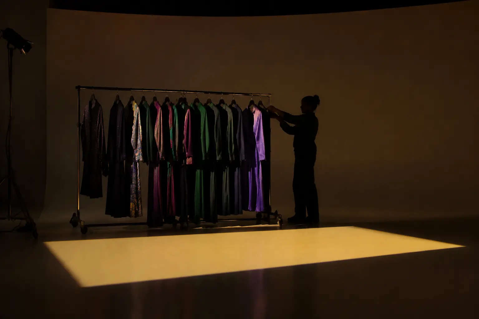 A stylist in silhouette adjusting a rail of original unbranded jewel-tone garments beside a rectangle of warm studio light